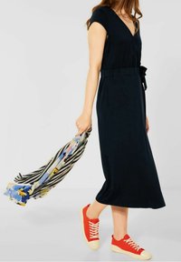 Granatowa sukienka do kolan z dekoltem w kształcie litery V i paskiem w talii. Noszona z czerwonymi trampkami. Trzyma w dłoni paski w kwiatowe akcenty.