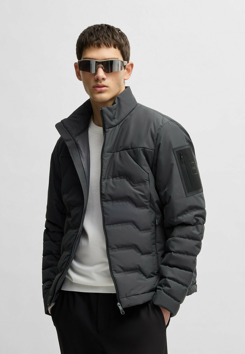 Grå puffjacka med hög krage, dragkedjeslutning och en dragkedjeficka på vänster ärm. Bärs med en vit skjorta under.