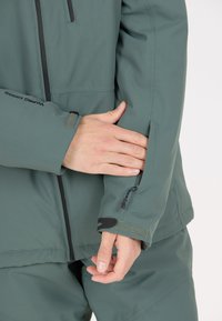 Grön isolerad jacka med en slät textur, åtsittande muddar och en ficka med dragkedja på vänster ärm. Logotyp på ärmen.