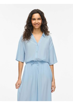 Femme aux longs cheveux bouclés souriante, portant une chemise ample bleu clair boutonnée et un pantalon large assorti avec une taille à cordon.