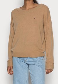 Beiger Strickpullover mit weitem Ausschnitt, überschnittenen Schultern, gerippten Bündchen und Saum, mit kleinem Logo auf der Brust. Kombiniert mit hellblauen Jeans.