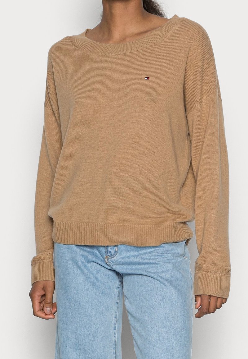 Beiger Strickpullover mit weitem Ausschnitt, überschnittenen Schultern, gerippten Bündchen und Saum, mit kleinem Logo auf der Brust. Kombiniert mit hellblauen Jeans.