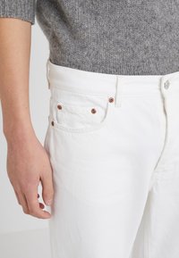 Vita denimjeans med en klassisk passform, fem fickor och nitar i kopparfärg. Texturen är slät med ett subtilt vävmönster.