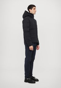 Black puffer jacket with a hood, featuring a quilted design. Worn over dark blue pants and black shoes. Simple, functional silhouette.