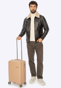 Bruine leren bomberjack met een beige schapenvachtkraag, houdend een tan hardshell koffer met diagonaal geribd patroon en uitschuifbaar handvat.