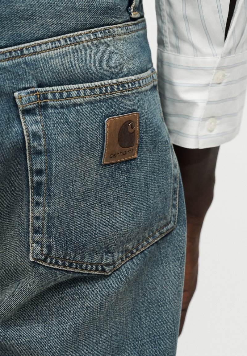 Gros plan sur la poche arrière en jean bleu avec un patch logo en cuir Carhartt et la main d'une personne portant une manche de chemise blanche rayée.