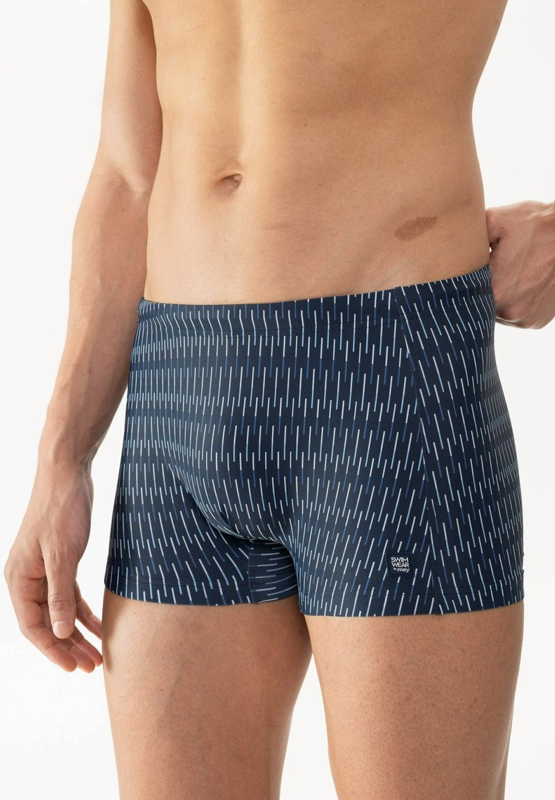 Navyblå badbyxor med vertikala vita ränder, tillverkade av elastiskt material. Har en figurnära design och en liten logotyp på midjebandet.