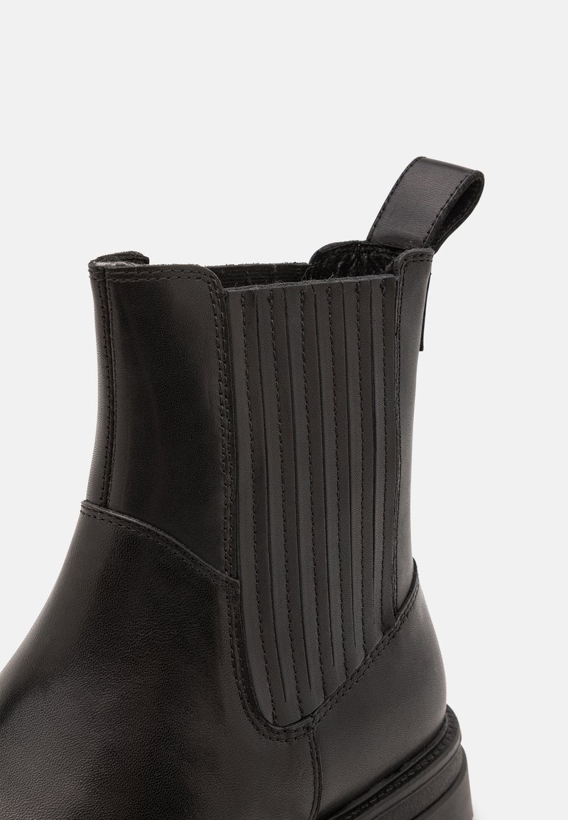 Stivale in pelle nera alla caviglia con un alto gambale, caratterizzato da accenti verticali a coste, una linguetta e una texture liscia. Dettaglio di suola robusta visibile.