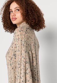 Suéter de malha em bege suave e texturizado com manchas pretas e pêssego; possui um corte solto e gola larga, exibindo padrões de costura intrincados.