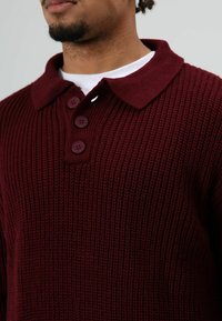 Man draagt een bordeauxrode gebreide trui met een kraag en drie knopen, over een wit ondershirt, zichtbaar van nek tot borst.