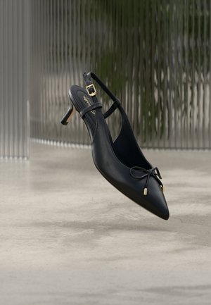 Chaussure à talon kitten et bride arrière pointue noire avec un petit nœud et une boucle dorée, présentée sur un sol en béton uni et un arrière-plan texturé.