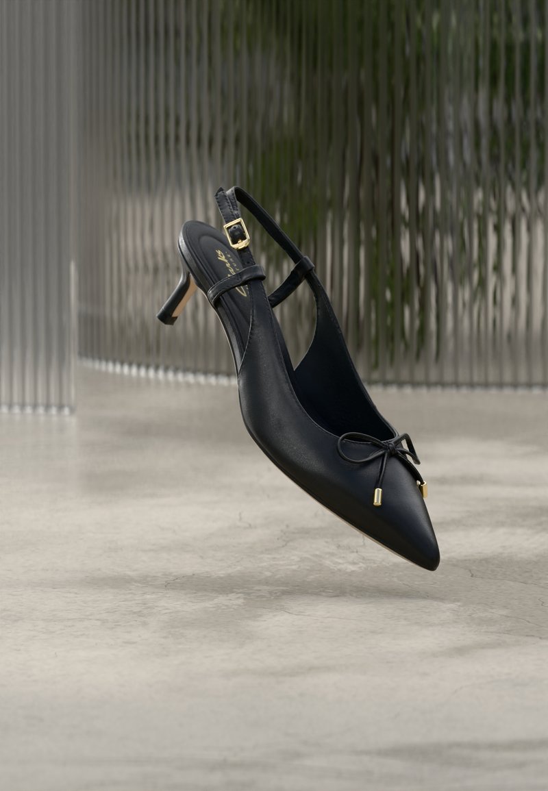 Μαύρο μυτερό μπρος-πίσω παπούτσι με χαμηλό τακούνι kitten, με μικρό φιόγκο και χρυσή αγκράφα, τοποθετημένο σε απλό δάπεδο από σκυρόδεμα και υφή φόντου.