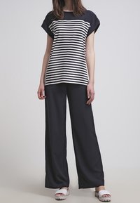 Woman wearing a black and white striped short-sleeve top, wide-leg black pants, and white slide sandals, standing on a gray floor.