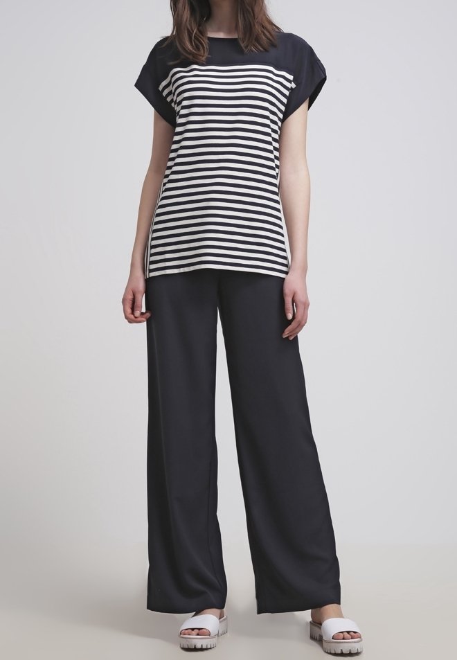 Woman wearing a black and white striped short-sleeve top, wide-leg black pants, and white slide sandals, standing on a gray floor.