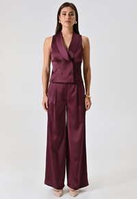 Completo in satin bordeaux composto da una parte superiore smanicata a doppio petto e pantaloni a gamba larga. Texture liscia con una vestibilità su misura.