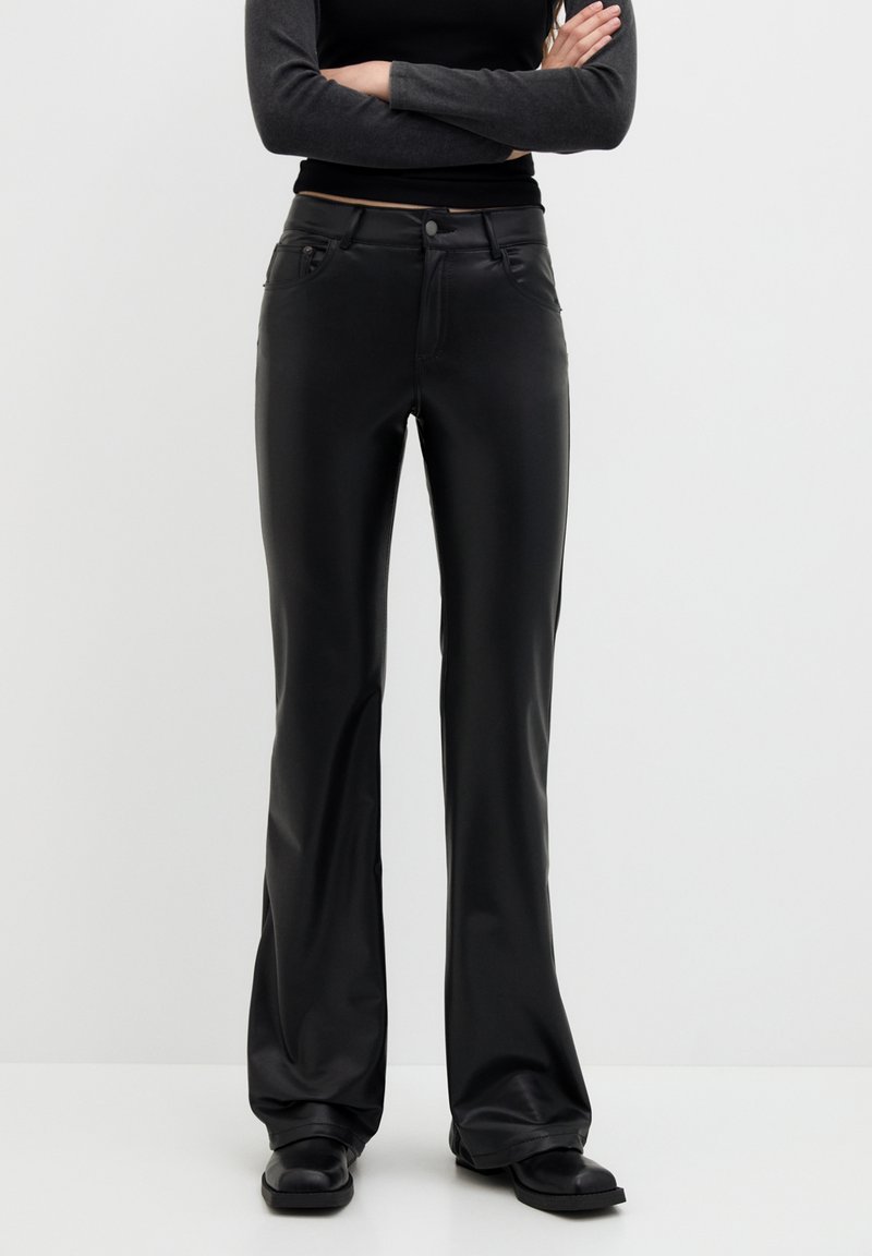 Pantalons évasés en cuir synthétique noir avec une fermeture éclair à l'avant, des poches et un léger éclat. Texture lisse avec des coutures contrastantes et une taille cintrée.