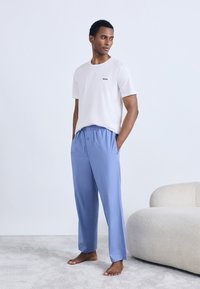 Maglietta bianca a maniche corte con logo "BOSS" abbinata a pantaloni larghi color azzurro chiaro, entrambi realizzati in tessuto morbido. Il modello è in piedi a piedi nudi.