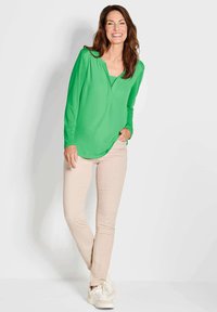 Langärmlige grüne Bluse mit V-Ausschnitt und abgerundetem Saum, kombiniert mit beigen Slim-Fit-Hosen und weißen Sneakern, vor einem neutralen Hintergrund stehend.