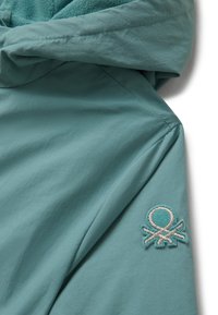 Detailní záběr rukávu a kapuce tyrkysové bundy s vyšitým logem značky na rukávu, ukazující texturu látky a detaily švů.