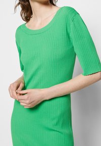 Robe verte en maille côtelée avec un col rond et des manches courtes, présentant une finition texturée et des bords de manches festonnés.