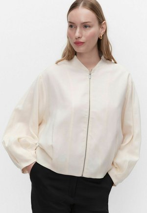 Kvinde iført en cremefarvet lynlåstrøje med lange ærmer og sorte bukser, stående med hænderne i lommerne og kigger let mod højre.