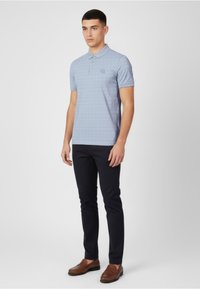 Polo azul claro con un patrón texturizado, tapeta de dos botones y logo bordado en el pecho. Combinado con pantalones oscuros y zapatos marrones.