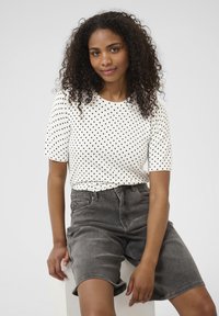 T-shirt blanc à pois noirs avec des manches courtes, associé à un short en denim gris. Le tissu semble doux avec une coupe décontractée. Design simple sans accents visibles.