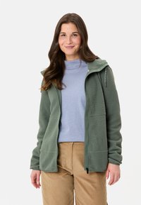 Giacca in pile con zip di colore verde chiaro, dotata di cappuccio, tasche con zip e tessuto testurizzato. Indossata sopra una maglietta azzurro chiaro e pantaloni in velluto a coste color beige.