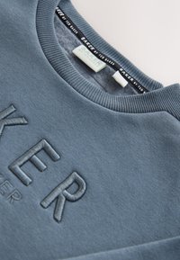 Hellblauer Sweatshirt mit rundem Ausschnitt, vorne mit erhabenen Buchstaben versehen. Hergestellt aus weichem, strukturiertem Stoff mit gerippten Bündchen.