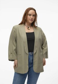 Blazer surdimensionné vert olive avec col châle, manches longues et poignets boutonnés, porté sur un haut noir et associé à un jean bleu.