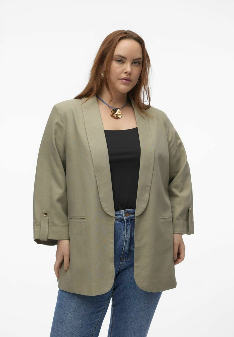 Blazer surdimensionné vert olive avec col châle, manches longues et poignets boutonnés, porté sur un haut noir et associé à un jean bleu.