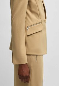 Beige bleiseri, joka on muotoiltu slim fit -malliin, sivussa vetoketjulla varustettu tasku, sileä pinta ja muotoillut hihat.