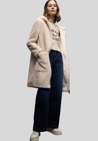 Płaszcz z futerka sztucznego w jasnym beżu z klapowym kołnierzem, bocznymi kieszeniami, noszony na beżowej bluzie i ciemnoniebieskich dzwonowatych dżinsach. Codzienne obuwie.