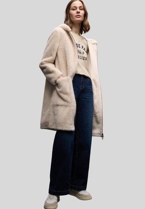 Płaszcz z futerka sztucznego w jasnym beżu z klapowym kołnierzem, bocznymi kieszeniami, noszony na beżowej bluzie i ciemnoniebieskich dzwonowatych dżinsach. Codzienne obuwie.