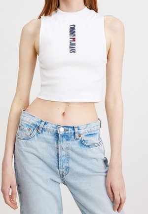 Personne mince portant un crop top blanc sans manches avec le logo Tommy Jeans et un jean en denim bleu clair taille haute avec bouton et fermeture éclair.