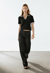 Černý pletený top s bílým límečkem a lemy na rukávech, v kombinaci s tmavě modrými džíny s vysokým pasem. Modelka stojí na neutrálním pozadí.