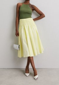 Débardeur vert côtelé associé à une jupe smockée jaune, accompagnée d'un sac à main argenté et de talons blancs pointus. Texture lisse, modèle mi-long.