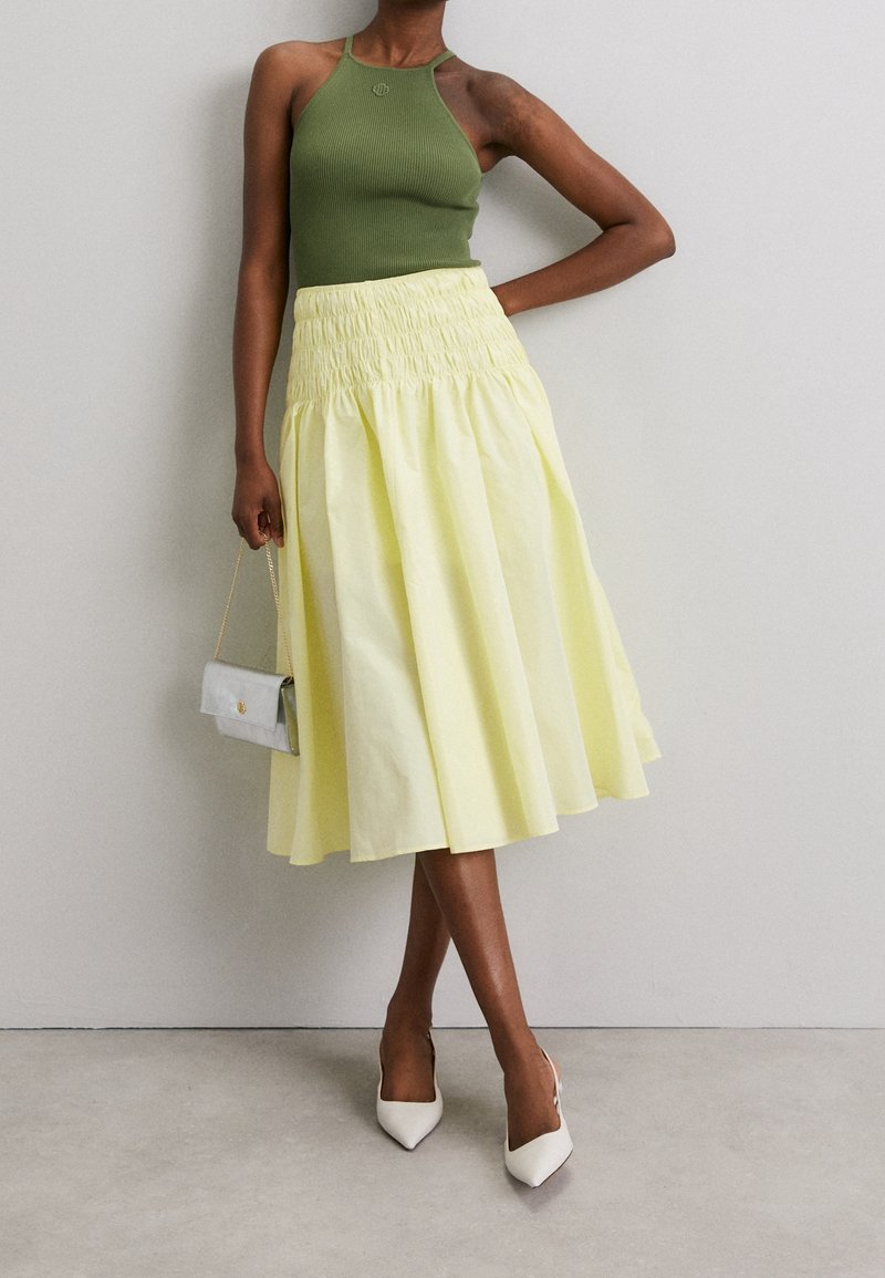 Débardeur vert côtelé associé à une jupe smockée jaune, accompagnée d'un sac à main argenté et de talons blancs pointus. Texture lisse, modèle mi-long.