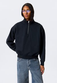 Svart genser med høy krage og glidelås foran. Myk tekstur, avslappet passform, og ribbestrikkede mansjetter. Brukt sammen med løse denimjeans.
