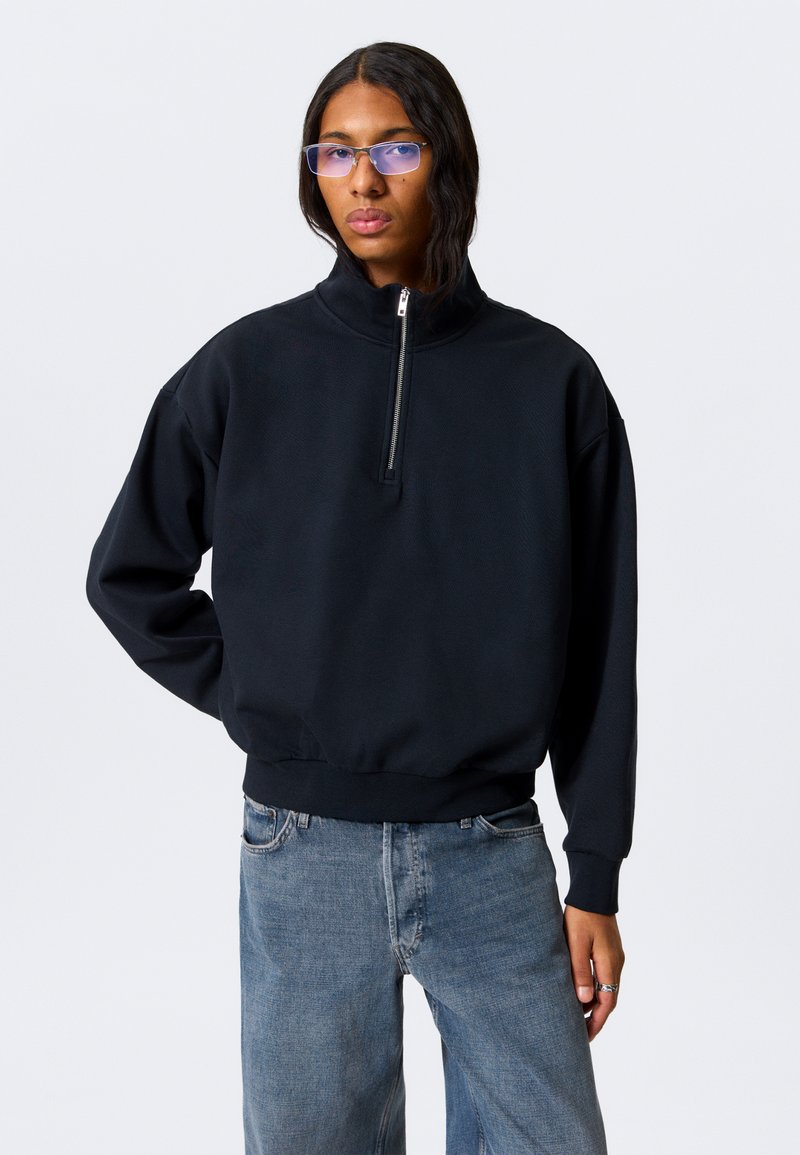 Svart genser med høy krage og glidelås foran. Myk tekstur, avslappet passform, og ribbestrikkede mansjetter. Brukt sammen med løse denimjeans.