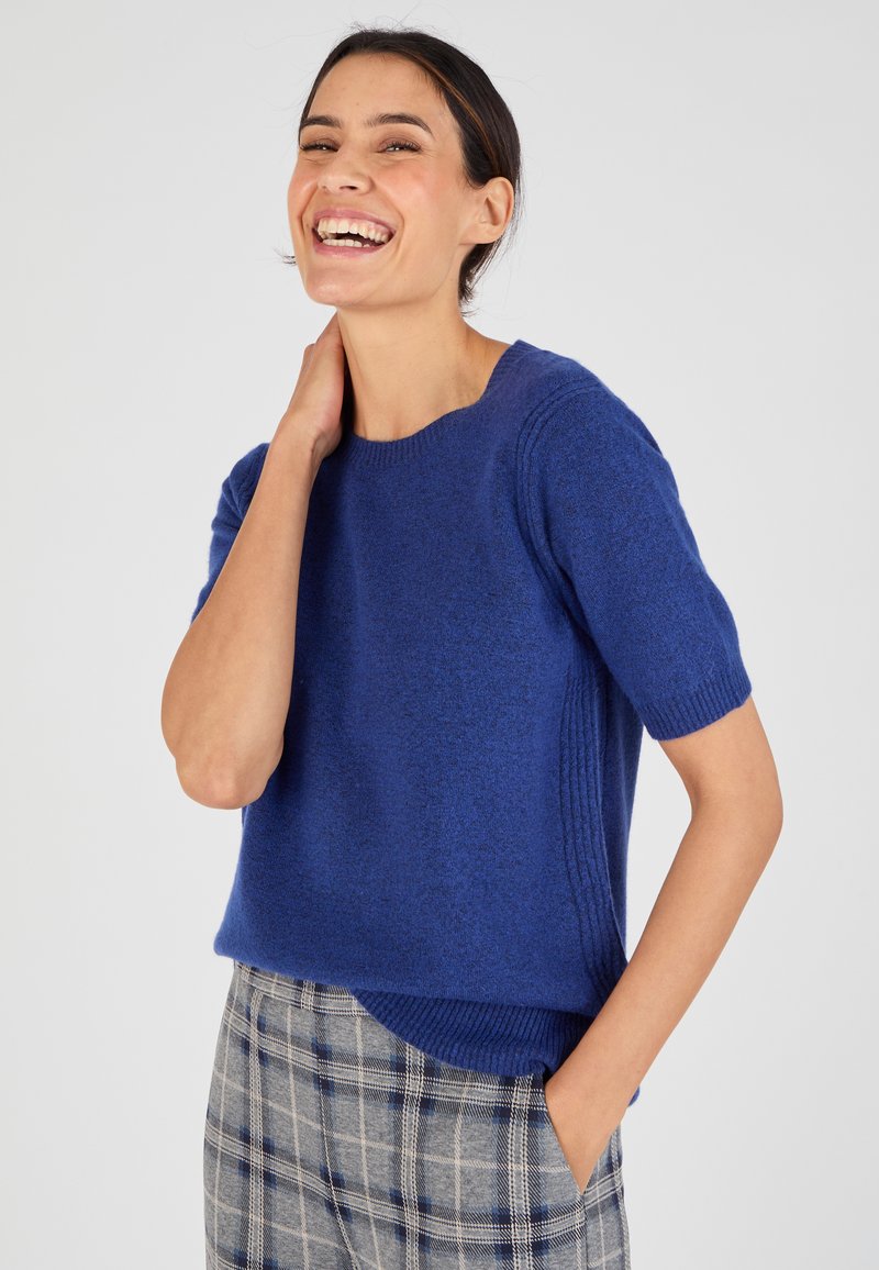 Pull bleu à manches courtes en matériau doux avec des accents côtelés sur les côtés, associé à un pantalon à carreaux gris.