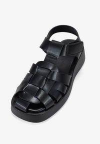 Sandalia de cuero trenzado negro con suela plana, puntera abierta y correa ajustable. Presenta un patrón texturizado y un diseño minimalista.