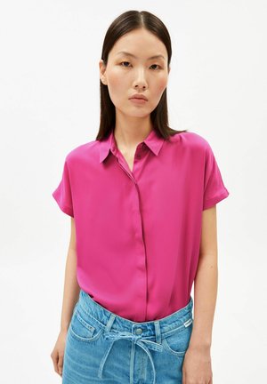 Vrouw met steil donker haar draagt een felroze blouse met korte mouwen, ingestopt in blauw, hooggetailleerd spijkerbroek met een ceintuur.