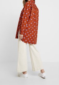 Kobieta w oversize'owej, rdzawopomarańczowej koszuli w lawendowe kropki, w szerokich kremowych spodniach i białych sneakersach, stojąca na tle jednolitego tła.
