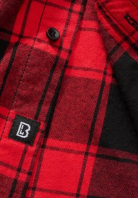 Camisa de franela a cuadros rojos y negros con una textura suave, cierre de botones y una etiqueta negra con un logo blanco en la esquina inferior izquierda.