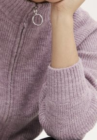 Fialový pletený svetr s vysokým límcem, který má stříbrný zip a ribbované manžety. Měkká textura a mírně volný střih.
