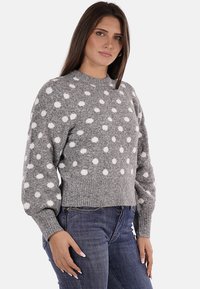 Maglione grigio lavorato a maglia con pois bianchi, scollatura rotonda e maniche lunghe. Il bordo a costine aggiunge texture. Indossato con jeans blu scuro.