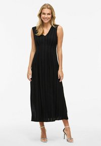 Abito maxi senza maniche di colore nero con pieghe verticali, scollatura a V e tessuto morbido. Indossato con tacchi a spillo argentati, che mettono in risalto il dettaglio della caviglia.