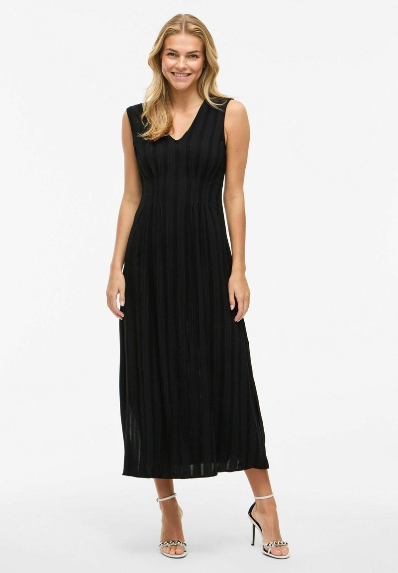 Abito maxi senza maniche di colore nero con pieghe verticali, scollatura a V e tessuto morbido. Indossato con tacchi a spillo argentati, che mettono in risalto il dettaglio della caviglia.