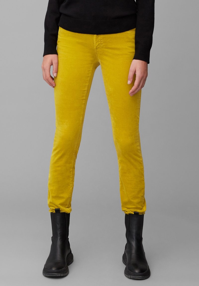 Pantalon en velours côtelé jaune à coupe slim, présentant une texture douce et une longueur cheville, associé à des bottines noires.
